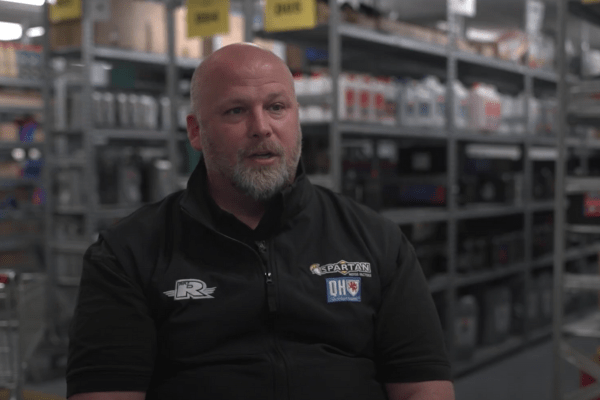 A man with a beard sits in a warehouse, wearing a black jacket with various logos. Shelves stocked with items are visible in the background.