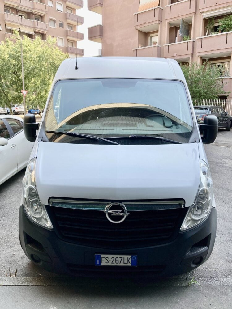 A white Opel van is parked on a street in front of apartment buildings, with a visible license plate reading "FS-267LK.