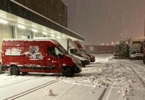 Red delivery vans parked in a snowy lot at night, with snow covering the ground and vehicles.