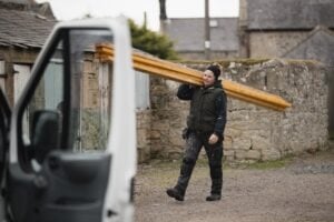 A person in workwear is carrying wooden beams on their shoulder, walking near a white van in a rural setting with stone buildings in the background.