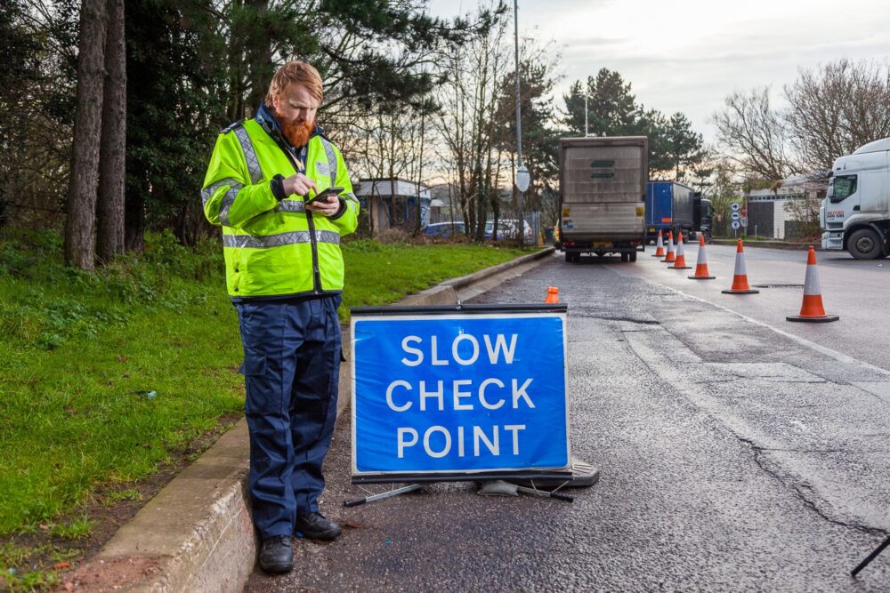 A person in a high-visibility jacket writes on a clipboard near a "Slow Check Point" sign on a roadside, with traffic cones and trucks in the background.