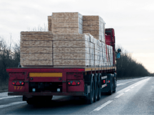 A red flatbed truck, with its load of plywood sheets expertly secured, travels down a road lined with bare trees beneath an overcast sky.