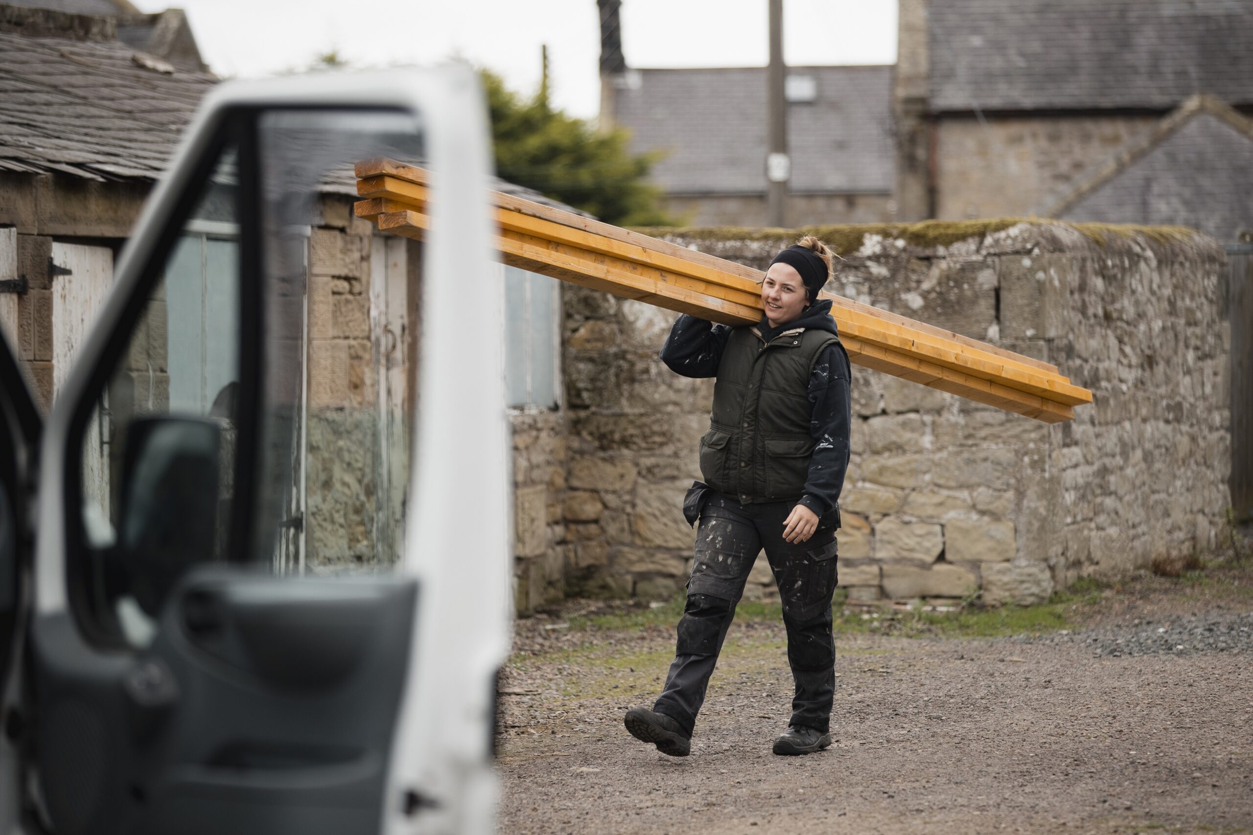 A person carries wooden planks on their shoulder in a rural setting, with stone buildings in the background and a white vehicle in the foreground.