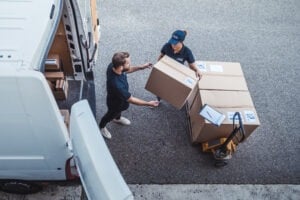 Two people in uniform load large cardboard boxes from a hand truck into a van parked on a paved surface. A clipboard and more boxes are visible in the van.