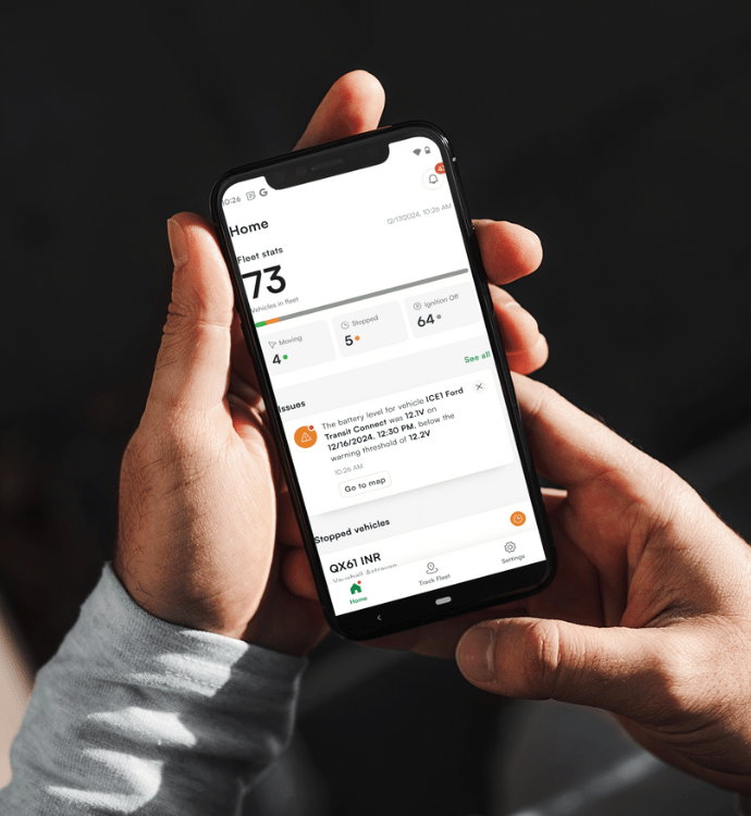 Person holding a smartphone displaying a fleet management app interface with vehicle statistics and notifications.