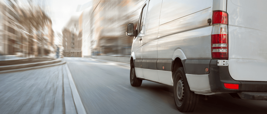 A white van speeds down a city street, seamlessly captured through vehicle tracking on the go, with motion blur emphasizing its rapid pace and the surrounding buildings slightly blurred in its wake.