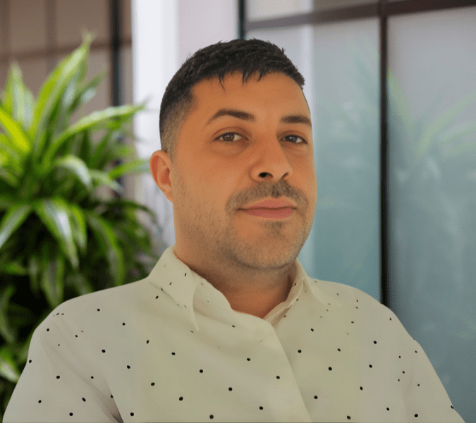 A person with short dark hair and a trimmed beard wears a white polka dot shirt, standing indoors with green plants and windows in the background.