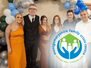 A group of six people stands together, with some dressed formally, as they support the MFCC charity. The logo of Montgomershire Family Crisis Centre is prominently displayed in front, while colorful balloons set a festive background.