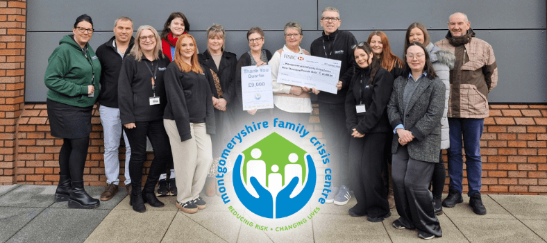 A group of people stands in front of a brick wall, proudly holding a large £10,000 check. They are smiling in their casual clothes, with the MFCC charity logo for Montgomeryshire Family Crisis Centre prominently displayed.