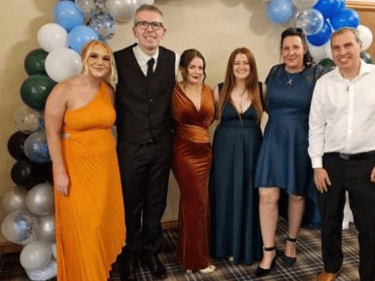 Six people in formal attire stand together in front of a balloon arch adorned with blue, green, black, and white balloons. Their poised gathering is part of the MFCC charity event, celebrating unity and generosity.