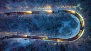 Aerial view of a winding, illuminated road through a snow-covered forest at night, with car headlights and taillights visible.