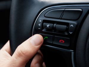 A hand pressing the green phone button on a car steering wheel control panel, with additional buttons for audio and call functions.