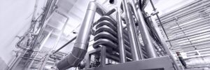 Industrial piping system with interconnected metal pipes and tubes, including a coiled section, inside a large facility with a high ceiling.